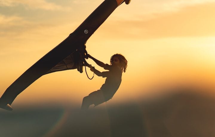 Wing foiler silhouetted against a golden sunset sky, holding the wing overhead