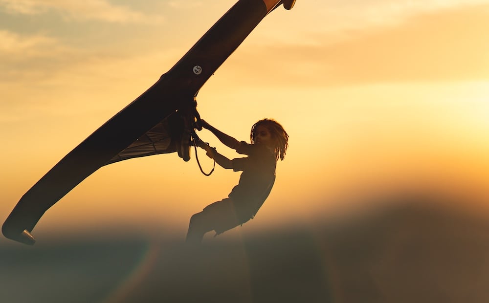 Wing foiler silhouetted against a golden sunset sky, holding the wing overhead